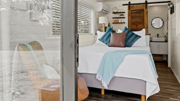 A bedroom with a bed, a wooden floor, a window, and a bathroom visible through an open door. - Container home | Roof Patio | Countryside Views (Fredericksburg)