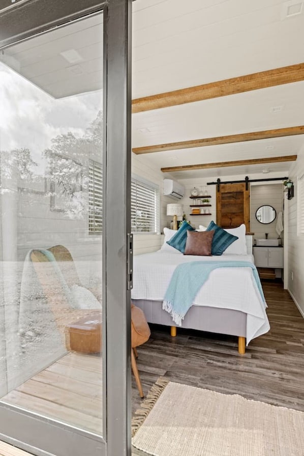 A bedroom with a bed, a wooden floor, a window, and a bathroom visible through an open door.