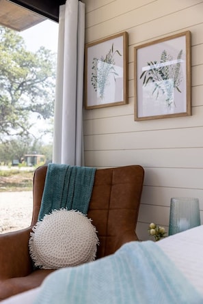 Una habitación con un sillón de cuero marrón, una manta color turquesa, un cojín blanco con textura e impresión enmarcadas de motivos botánicos en la pared.
