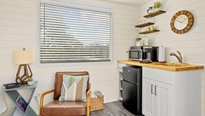A compact kitchen with a built-in microwave, a sink, and a refrigerator. There is a window with blinds, a chair with a cushion, and a lamp on a side table. - New! The Sunset Luxury Container Home (Fredericksburg)
