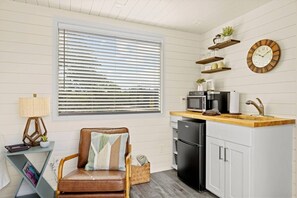 A compact kitchen with a built-in microwave, a sink, and a refrigerator. There is a window with blinds, a chair with a cushion, and a lamp on a side table. - New! The Sunset Luxury Container Home (Fredericksburg)