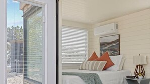 A bedroom with a bed, pillows, a bedside table, a lamp, and a window with blinds. - New! The Sunset Luxury Container Home (Fredericksburg)