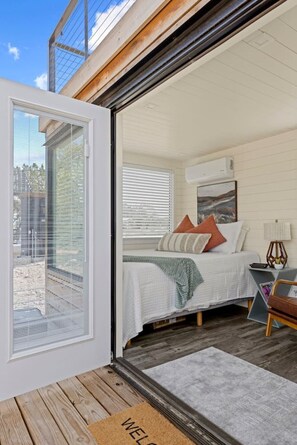 A bedroom with a bed, pillows, a bedside table, a lamp, and a window with blinds. - New! The Sunset Luxury Container Home (Fredericksburg)