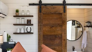 A modern bathroom with a wooden sliding door, shelves with glassware, a mirror, and a sink. - New! The Perry-Chic container home Oasis (Fredericksburg)