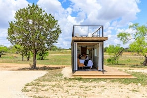 Una pequeña construcción al aire libre con una cama y un ventanal, rodeada de árboles y un cielo despejado.