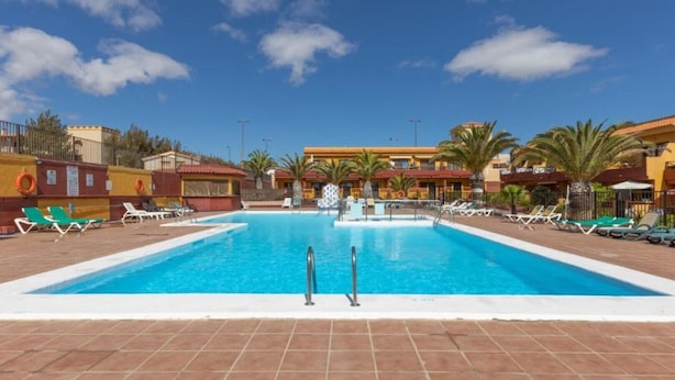 Una piscina con tumbonas y palmeras, rodeada de edificios con fachadas rojas y amarillas.
