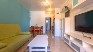 Una sala de estar con un sofá amarillo, una mesa de centro blanca y un televisor de pantalla plana sobre un soporte blanco.