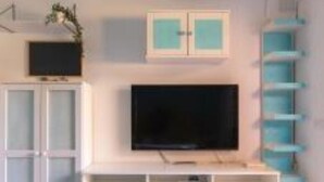 Una habitación con una televisión de pantalla plana sobre un soporte blanco, una planta pequeña y un mueble blanco con puertas de vidrio.