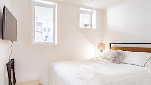 Un dormitorio con una cama, un televisor montado en la pared y dos ventanas.