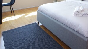 Ein ordentlich bezogenes Bett mit weißen Leinen und aufgerollten Handtüchern, ein Nachttisch mit Lampe und ein Sessel in einem modernen Zimmer mit Holzboden und einer grauen Akzentwand.