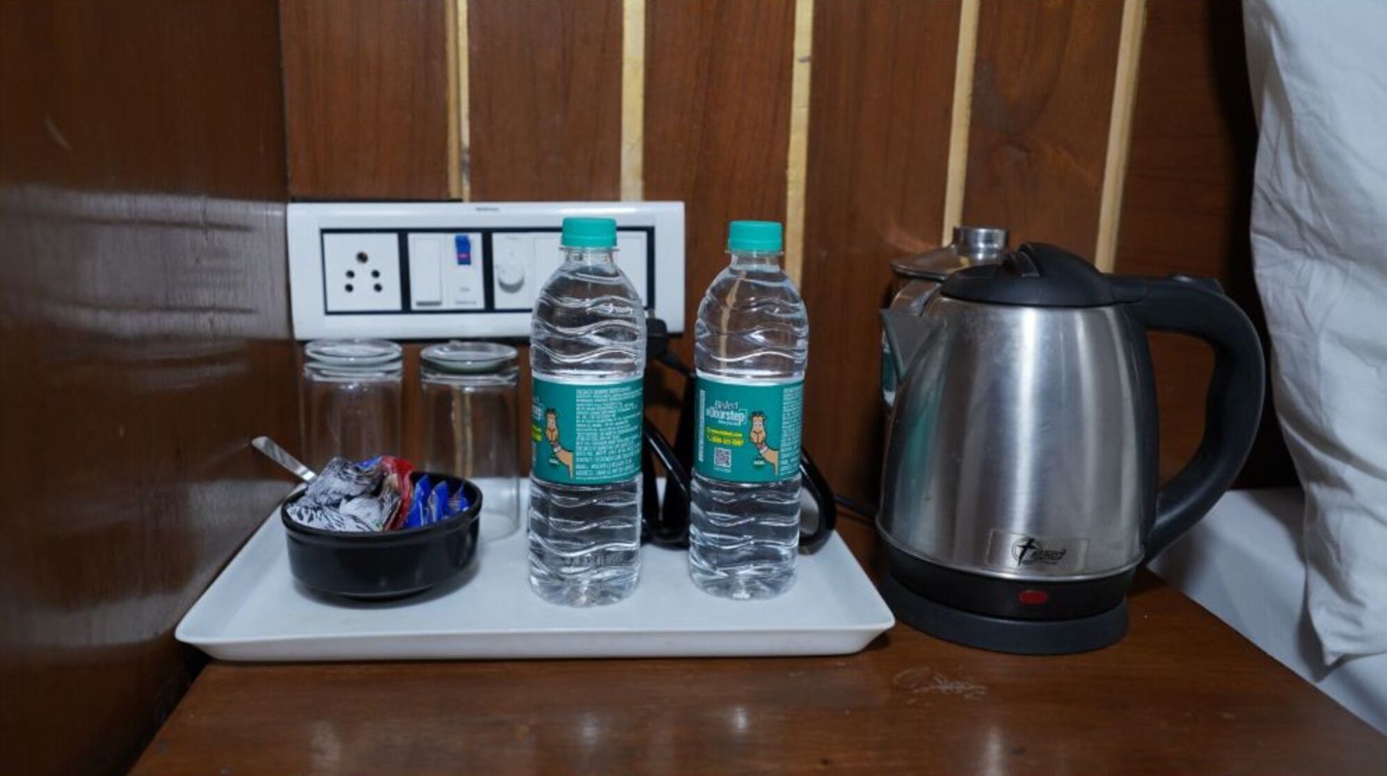 Habitación de hotel con una mesita blanca que sostiene un cuenco negro con cubiertos, dos botellas de agua y un hervidor de acero inoxidable.