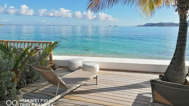 Een houten terras met een lounge stoel en kussens, met uitzicht op een kalme zee met boten en een eiland in de verte.