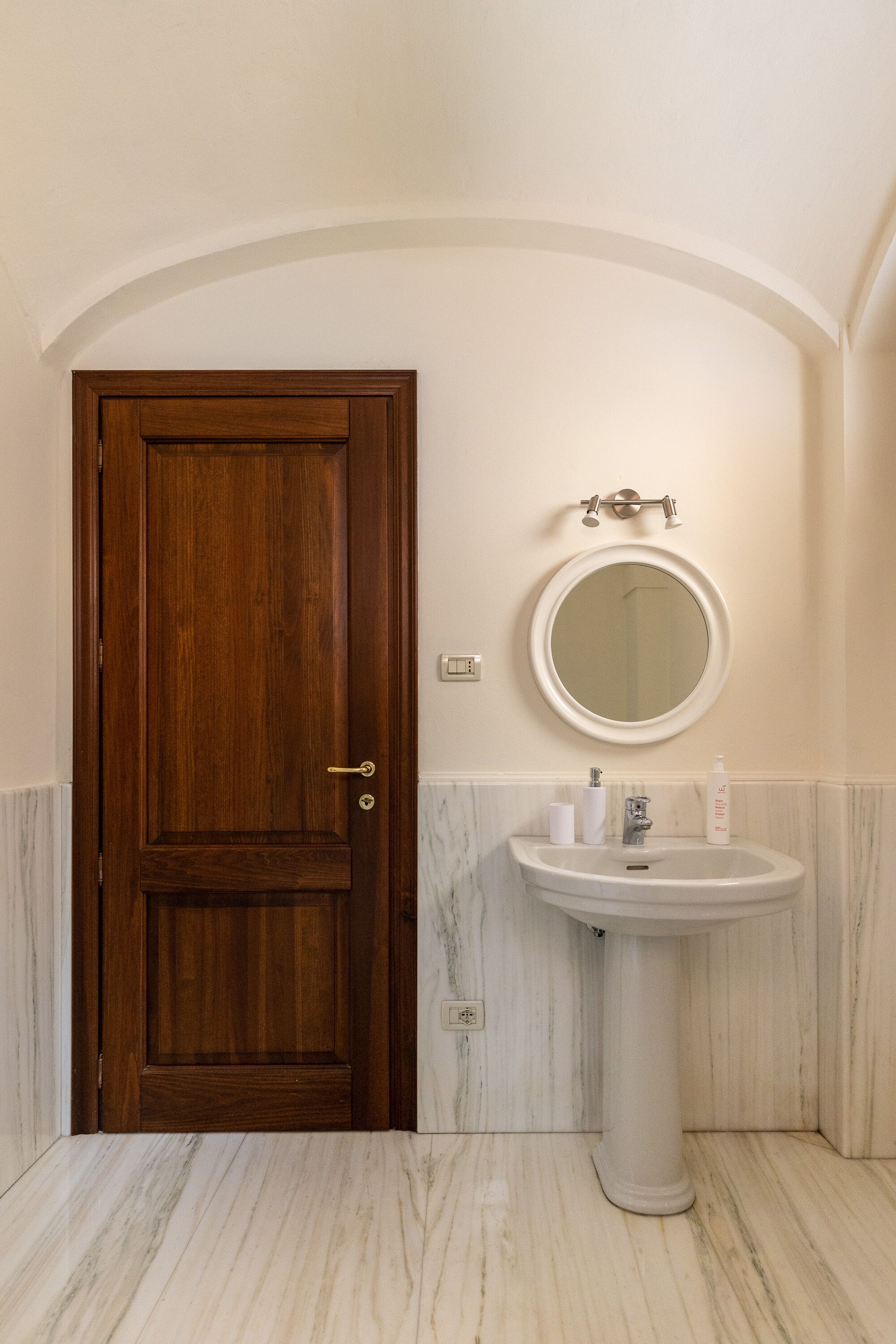 A bathroom with a wooden door, a round mirror, a pedestal sink, and marble flooring.