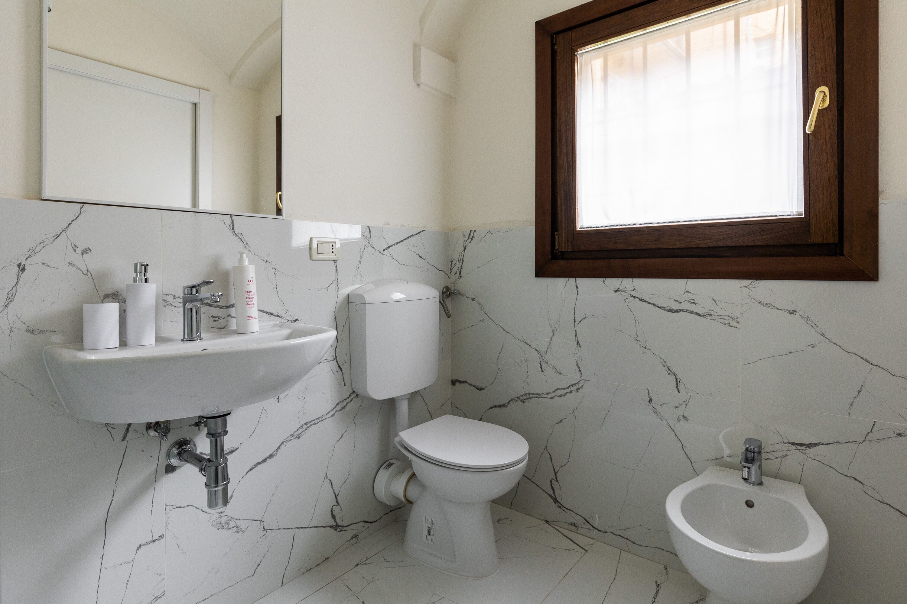 A bathroom with a marble sink, toilet, bidet, and a window with curtains.