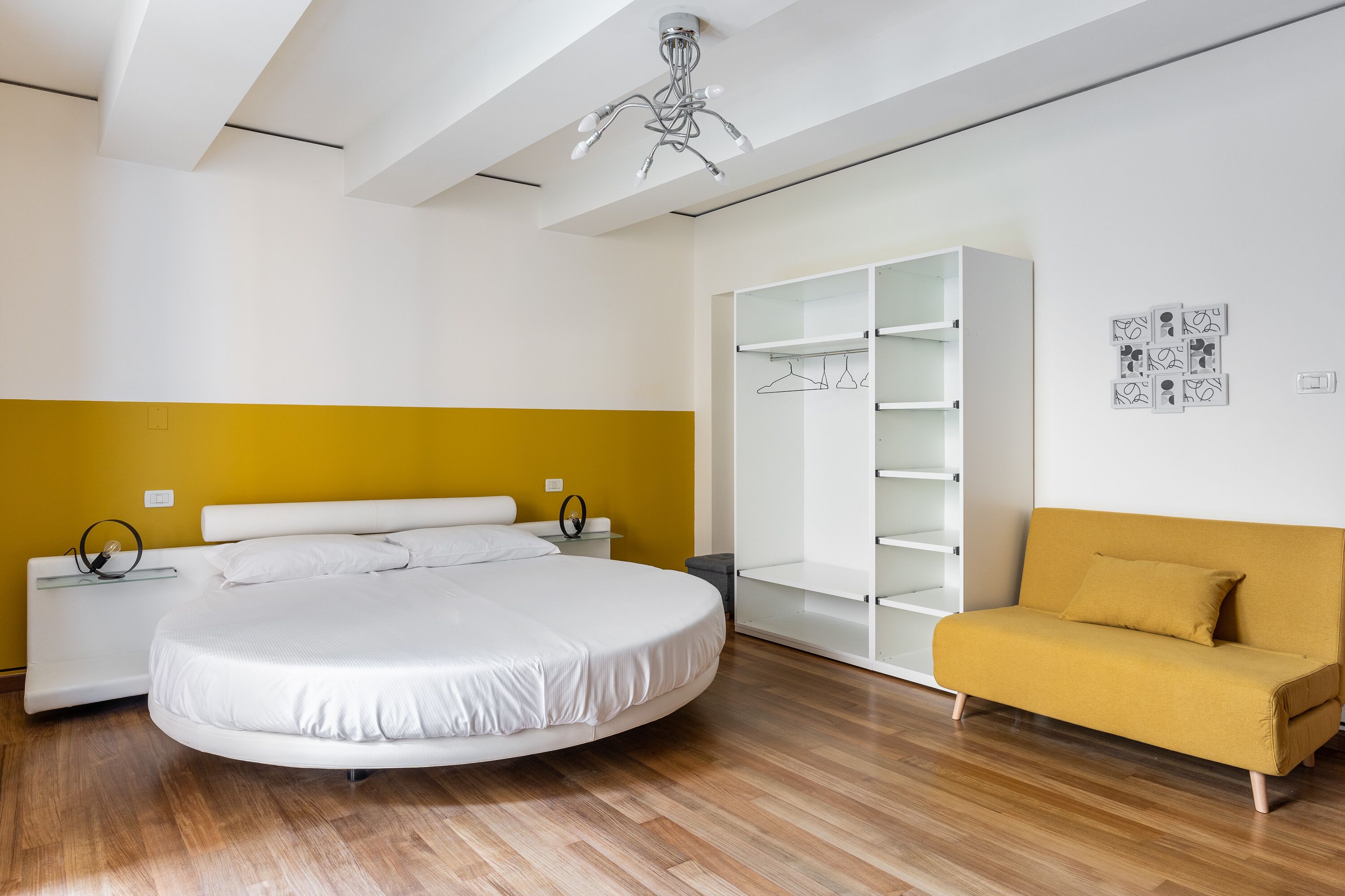 Ein modernes Schlafzimmer mit einem runden Bett, einem gelben Sofa, einem weißen Kleiderschrank und einer Deckenleuchte.
