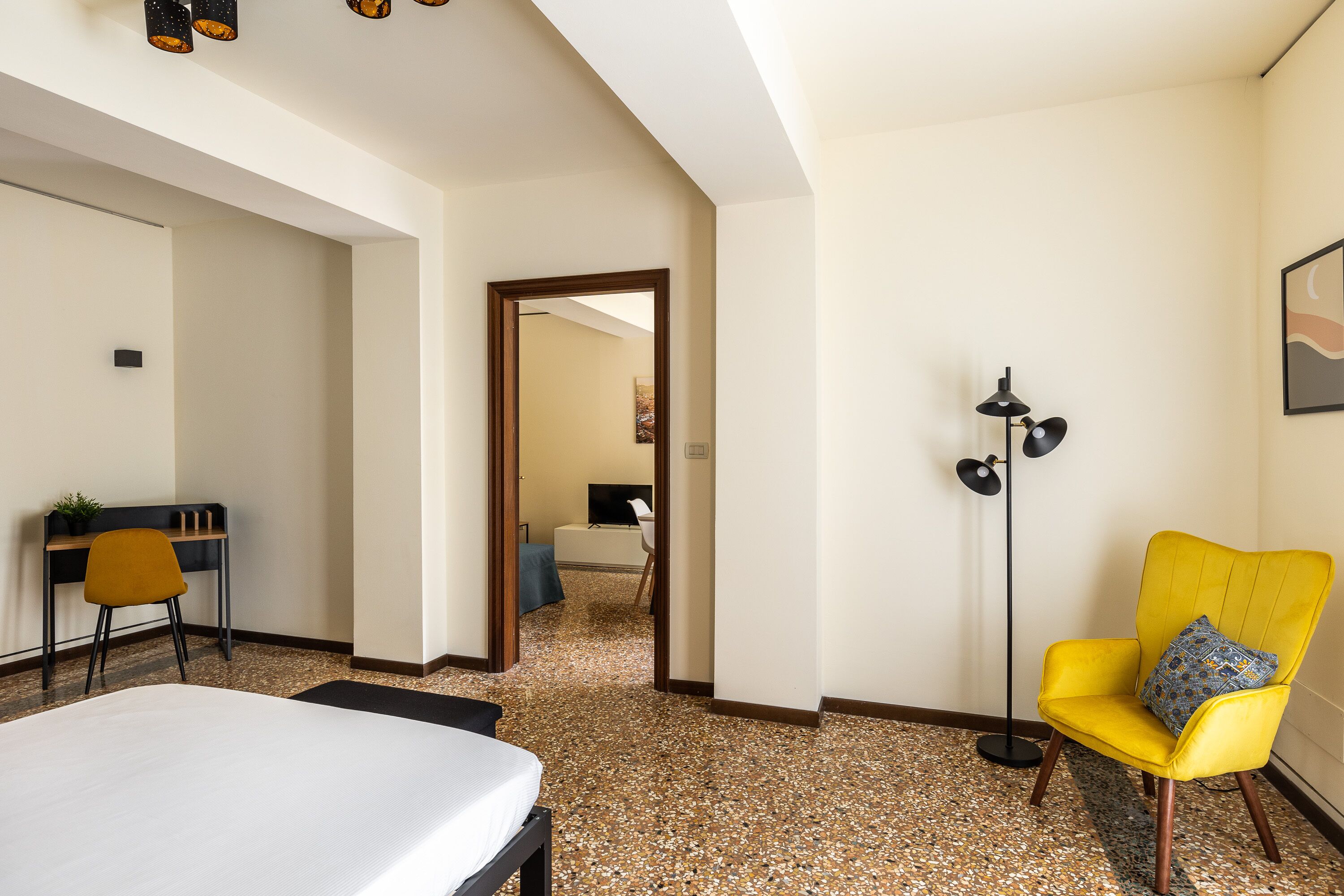 Una camera d'albergo con un letto, una scrivania con una sedia, una poltrona gialla, una lampada da terra e un quadro appeso al muro.