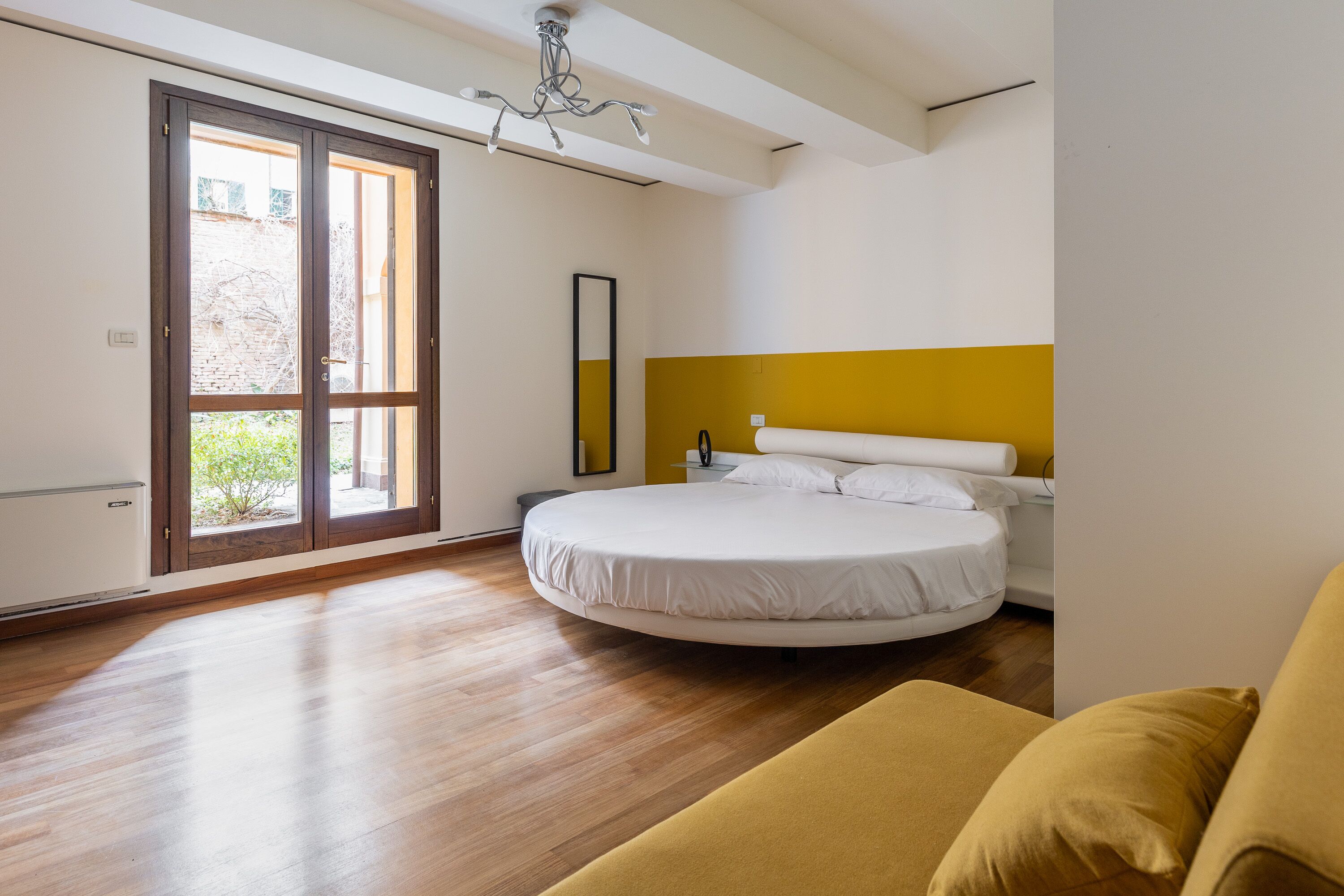 Une chambre à coucher avec un lit rond, un mur d’accent jaune et une grande fenêtre donnant sur un paysage verdoyant.