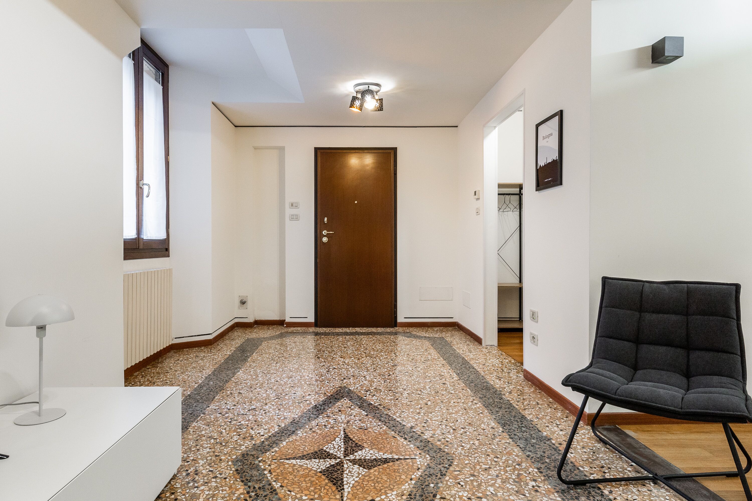 A hallway with a patterned floor, a wooden door, a black chair, and a white desk with a lamp.