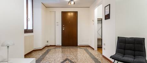 A hallway with a patterned floor, a wooden door, a black chair, and a white desk with a lamp.