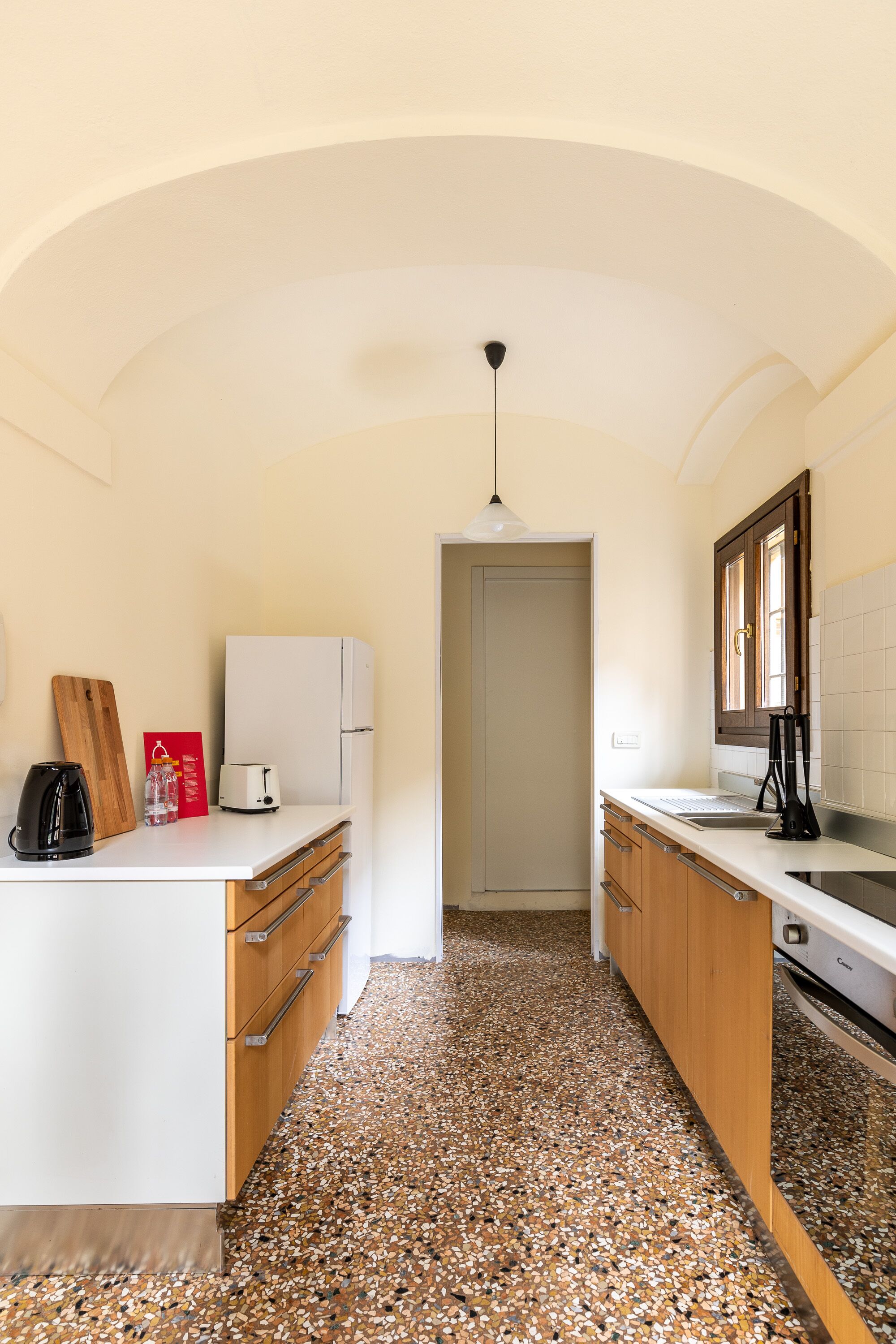A compact kitchen with a white refrigerator, wooden cabinets, a countertop with a kettle and a toaster, and a speckled tile floor.
