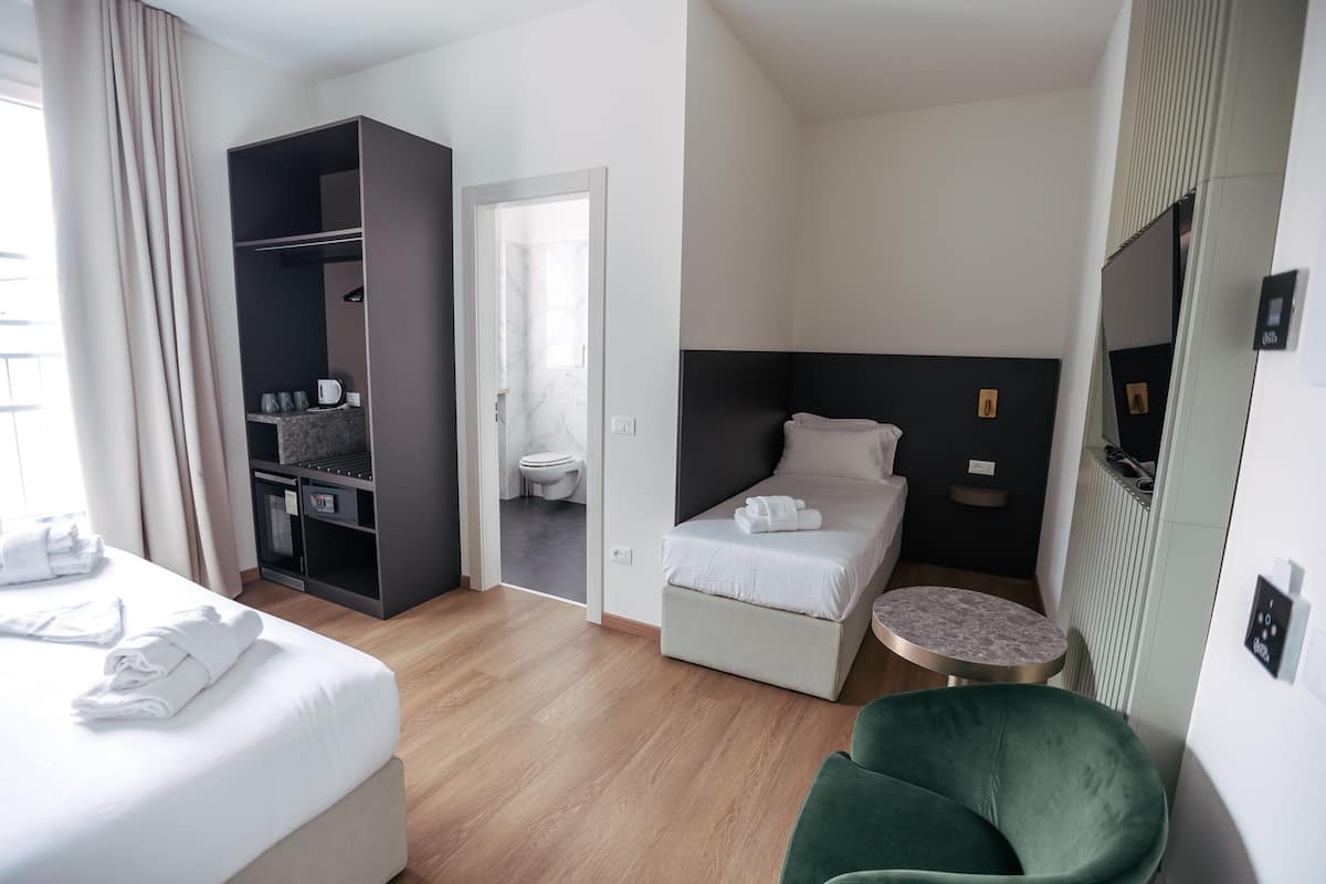 A hotel room with a bed, a green armchair, a small round table, a wardrobe, and a bathroom visible through an open door.
