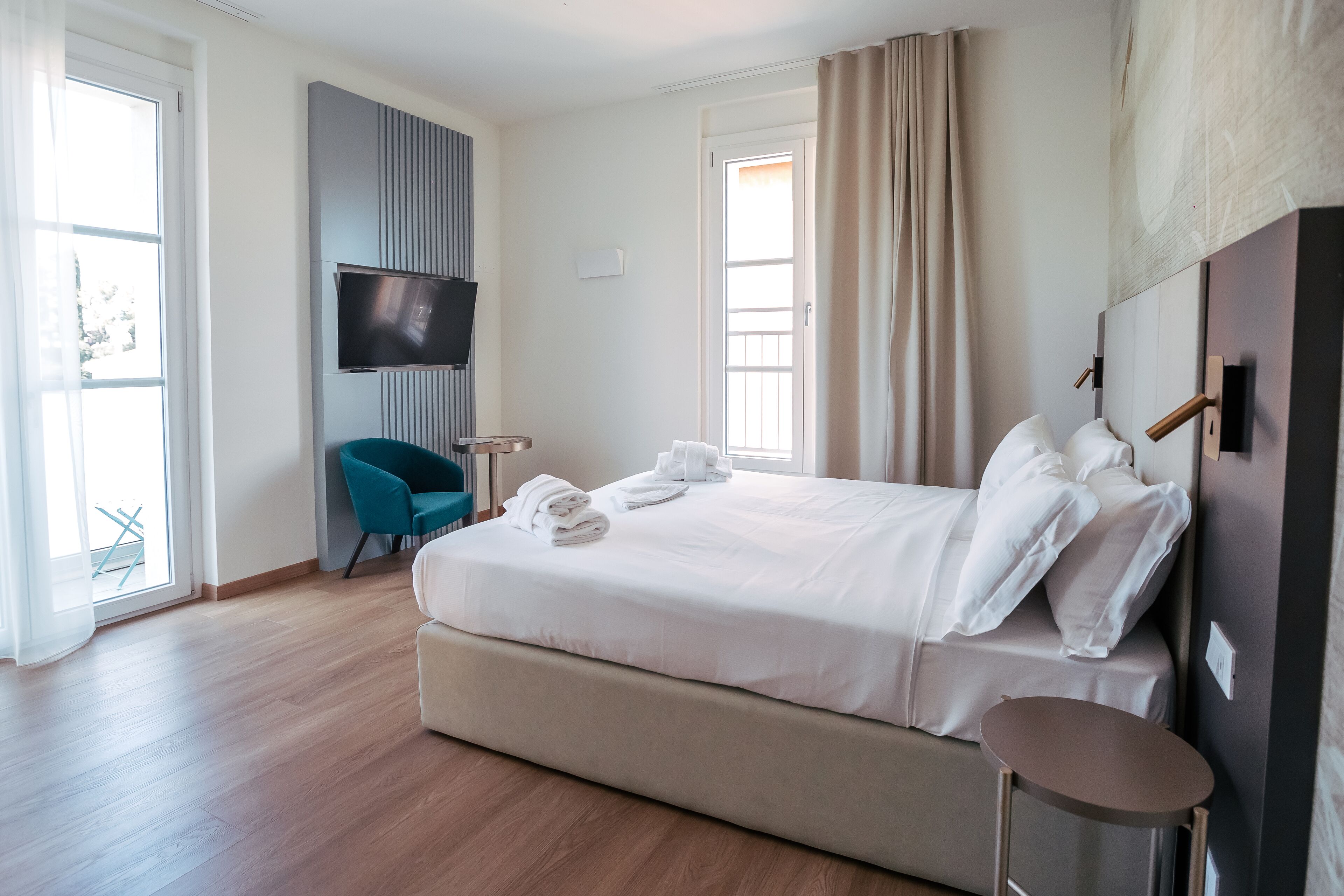 A modern hotel room with a large bed, a flat-screen TV mounted on the wall, a blue chair, and a small table.