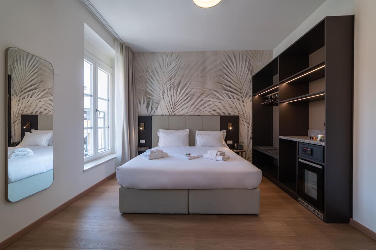 A modern hotel room with a large bed, a built-in wardrobe, a kitchen area, and a large mirror.