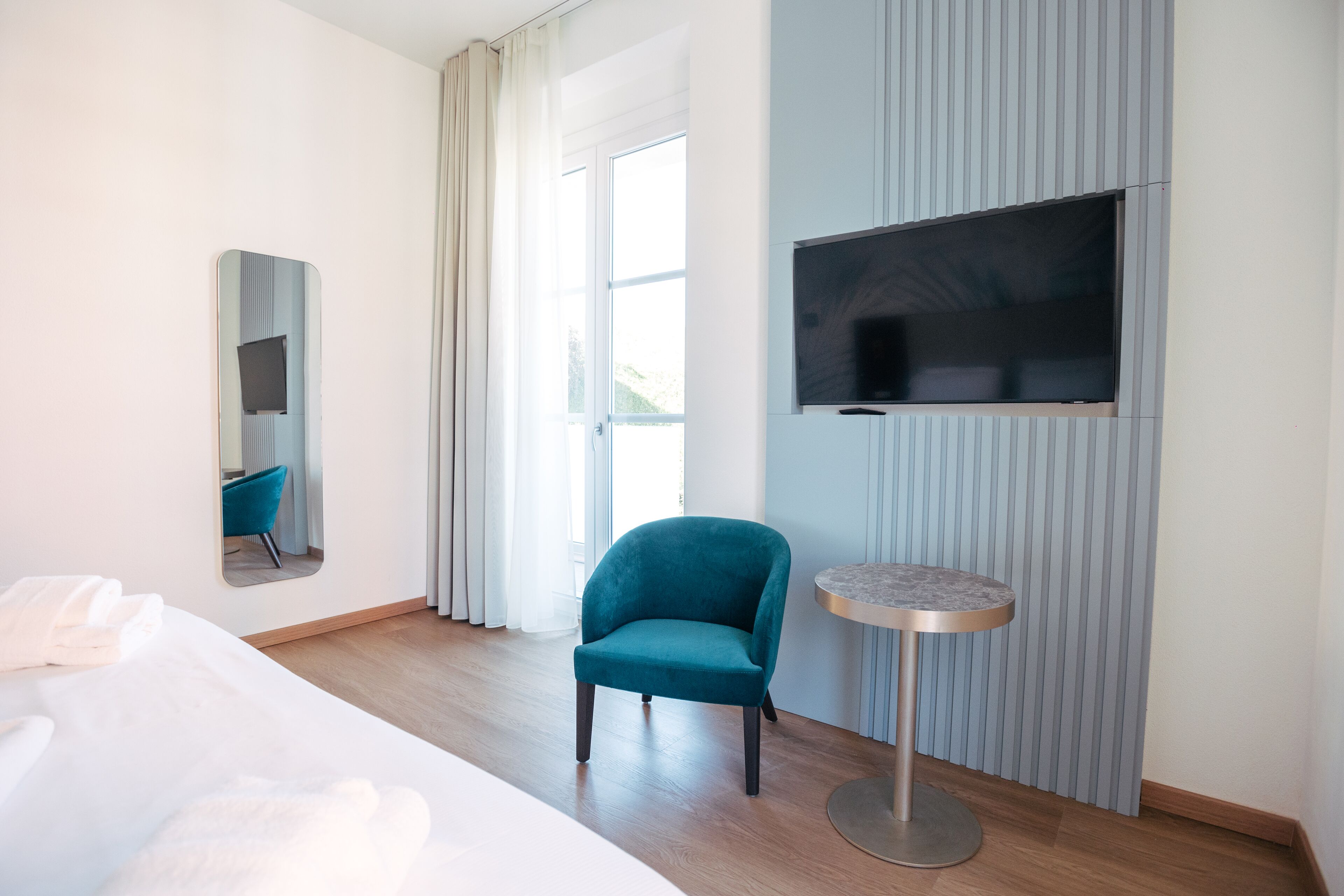 Ein Hotelzimmer mit einem Bett, einem türkisfarbenen Sessel, einem runden Tisch, einem an der Wand montierten Fernseher und einem großen Fenster mit Vorhängen.