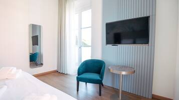 Ein Hotelzimmer mit einem Bett, einem türkisfarbenen Sessel, einem runden Tisch, einem an der Wand montierten Fernseher und einem großen Fenster mit Vorhängen.