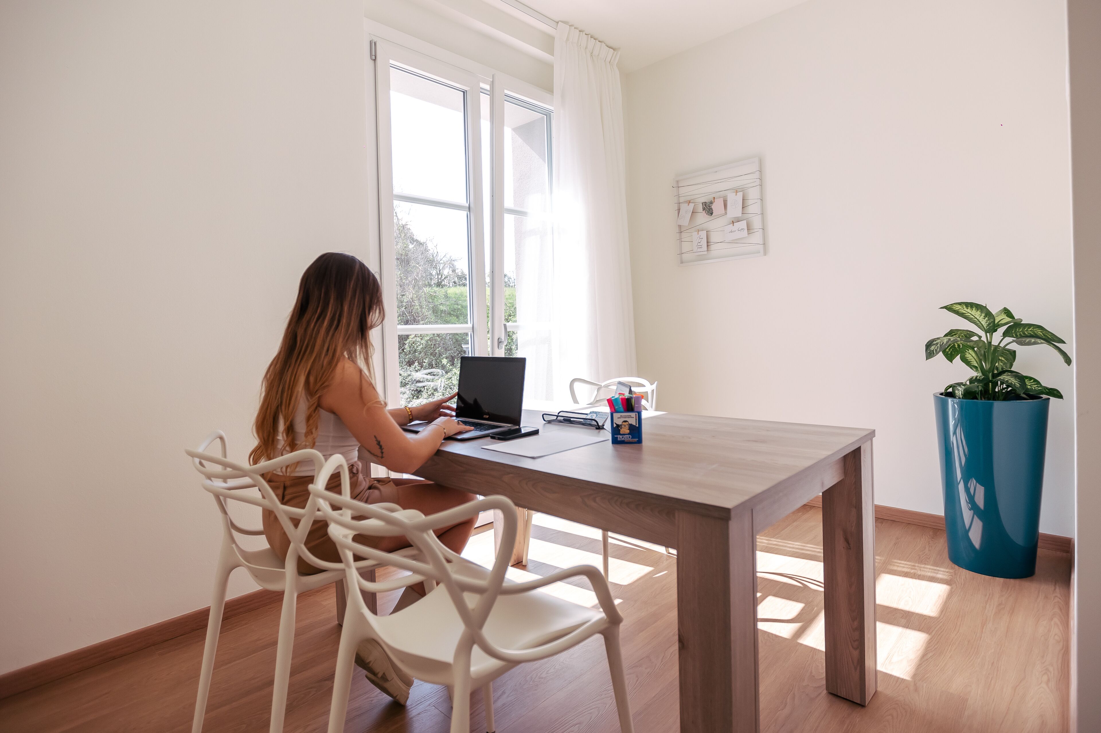A person working at a wooden desk with a laptop, white chairs, a potted plant, and a window with curtains.