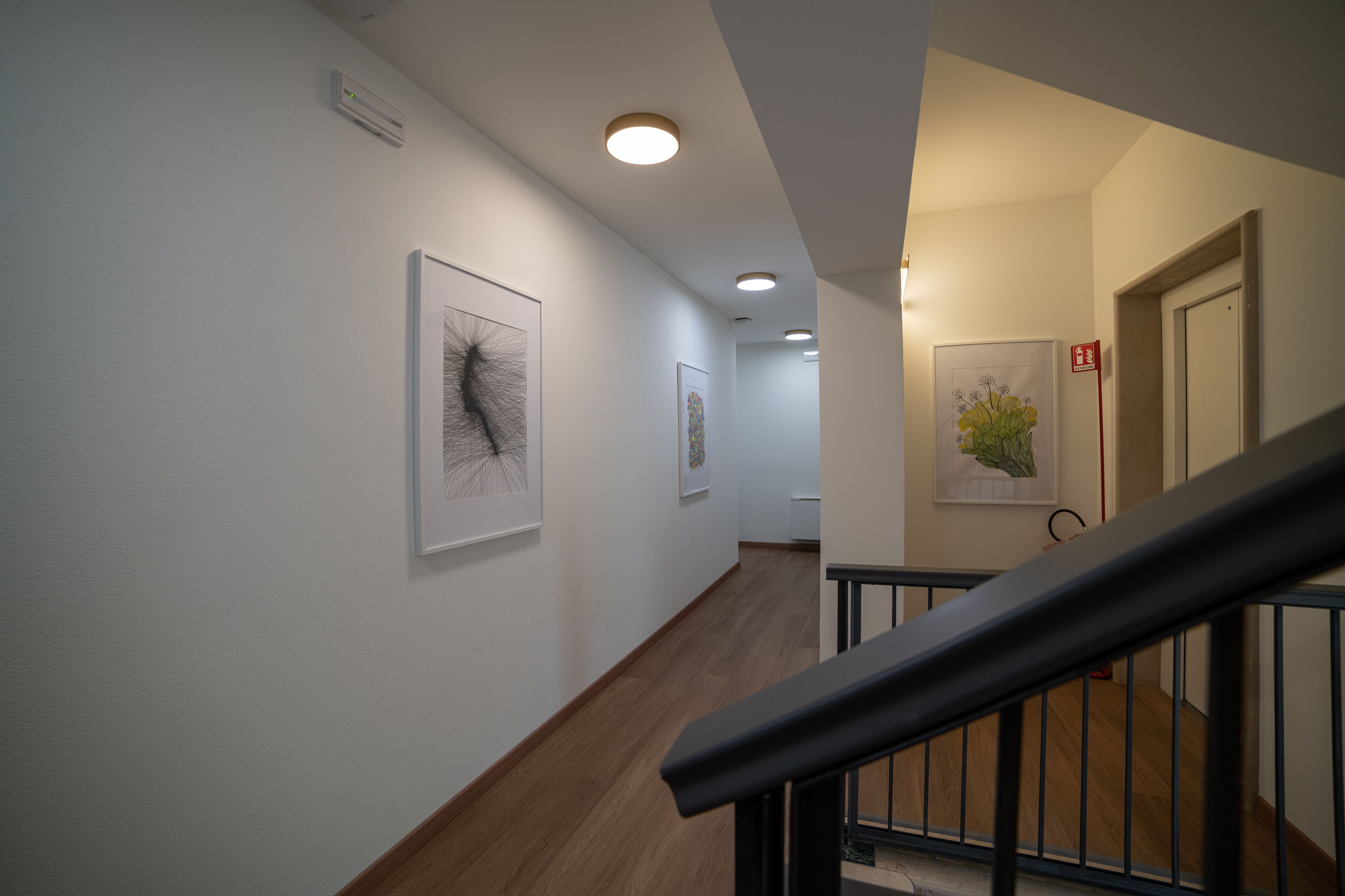 A hallway with framed artwork on the walls, a wooden floor, and a staircase with a metal railing.
