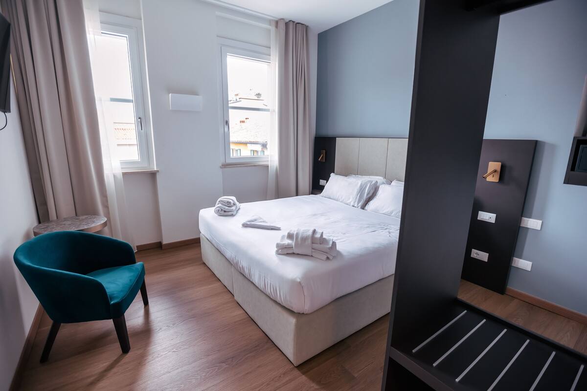 A hotel room with a bed, a teal chair, a small round table, and a view of the outside through a window.