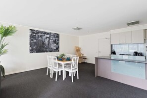 A modern kitchen with white cabinets and a glass countertop, a dining area with a table and chairs, and a black and white cityscape artwork on the wall.
