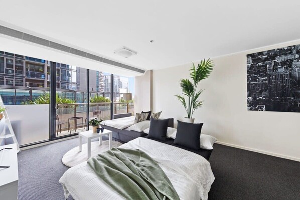 A modern hotel room with a large bed, a balcony with a city view, and a potted plant.