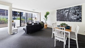 A modern living room with a black sofa, white dining table, and chairs. There is a large window with a cityscape view, a potted plant, and a black and white cityscape painting on the wall. - A Large 2BR Apt Near Crown, Sleeps 8, FREE Parking (Southbank)
