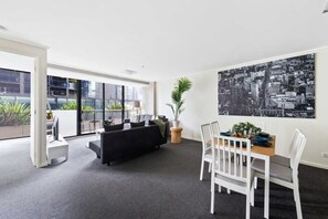 Una sala de estar moderna con un sofá negro, una mesa de comedor blanca y sillas. Hay un ventanal con vista a la ciudad, una planta en maceta y un cuadro de paisaje urbano en blanco y negro en la pared.