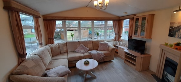 A cozy living room with a large L-shaped sofa, a small round coffee table, a flat-screen TV, and a fireplace. - Mobile home "Uns Fischernüst 2" - your cozy retreat in East Frisia (Hinte)