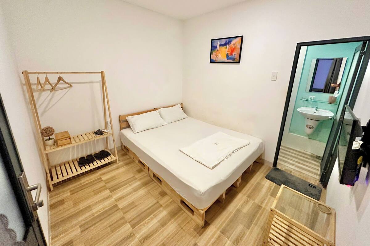 A small, clean room with a single bed, a wooden shoe rack, a small shelf with toiletries, and a bathroom visible through a glass door.