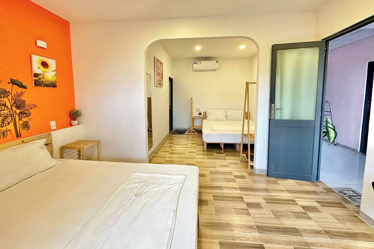 A hotel room with a bed, a wooden floor, a small table, a nightstand, a mirror, and a door leading to another room.