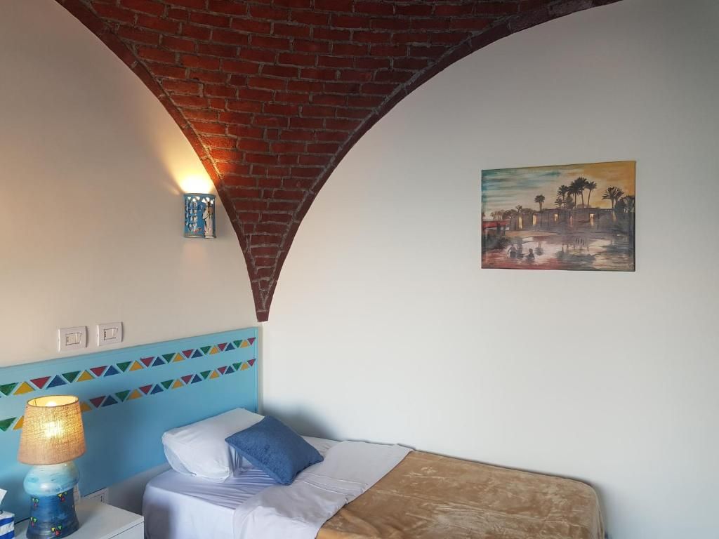 A hotel room with a brick arch ceiling, a bed with a blue and white headboard, a wall-mounted lamp, and a painting of a tropical scene.