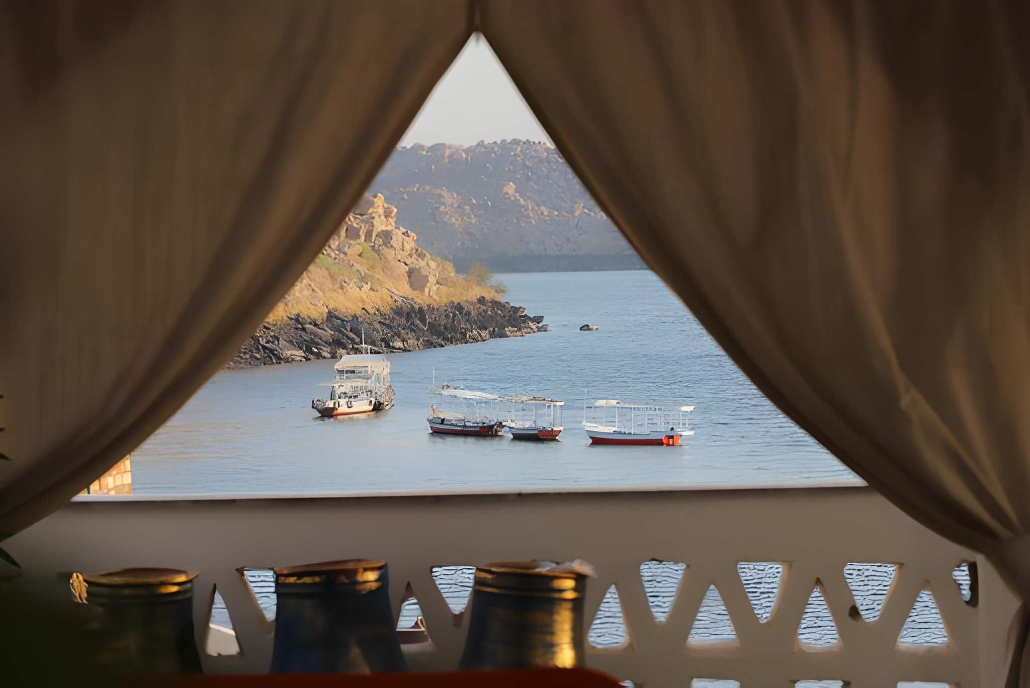 Vista desde una ventana con cortinas, sobre un cuerpo de agua tranquilo con varios barcos y un litoral rocoso.