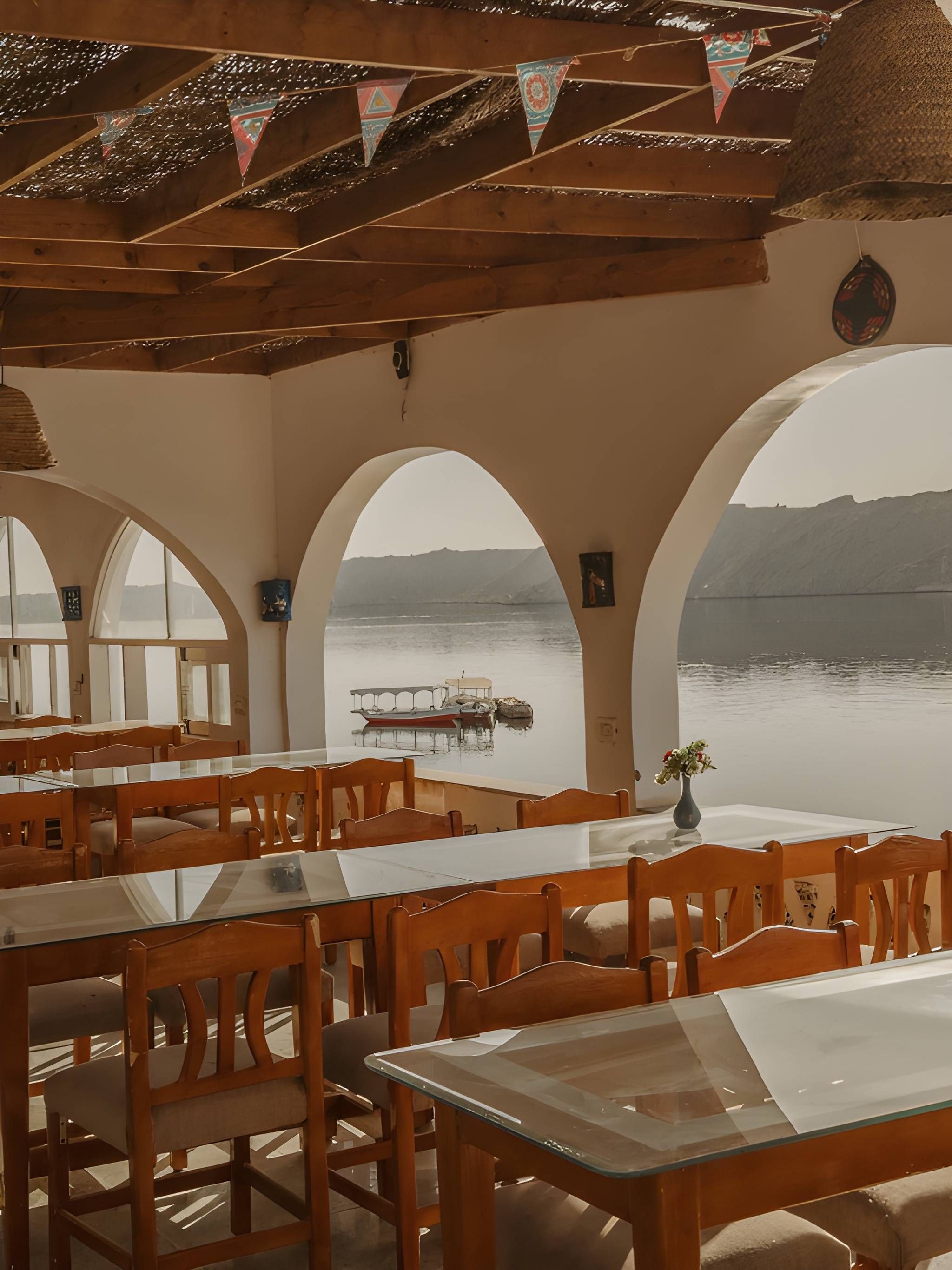 A restaurant with wooden tables and chairs, large arched windows, and a view of a lake with boats.
