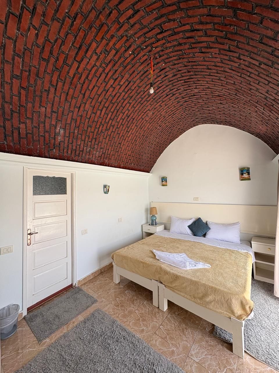 Quarto com teto em arco de tijolos, cama com cobertura bege, paredes brancas e pequena mesa de cabeceira com luminária.