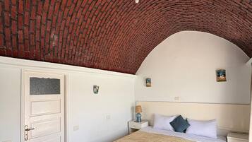 Quarto com teto em arco de tijolos, cama com cobertura bege, paredes brancas e pequena mesa de cabeceira com luminária.