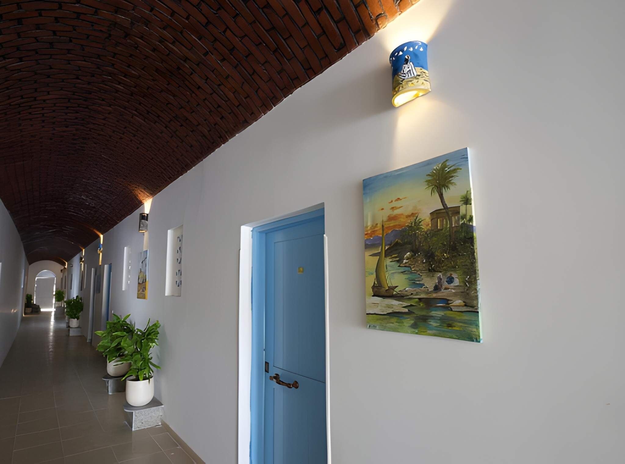 Un couloir avec une porte bleue, des plantes en pot et un tableau représentant un paysage.