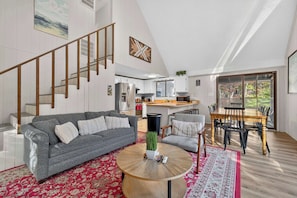 A spacious living area with a grey sofa, a wooden coffee table, and a dining table with chairs. A staircase with wooden railings leads to an upper level.