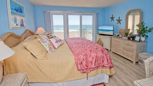 A bedroom with a large bed, a dresser, a television, and a view of the ocean through the window.