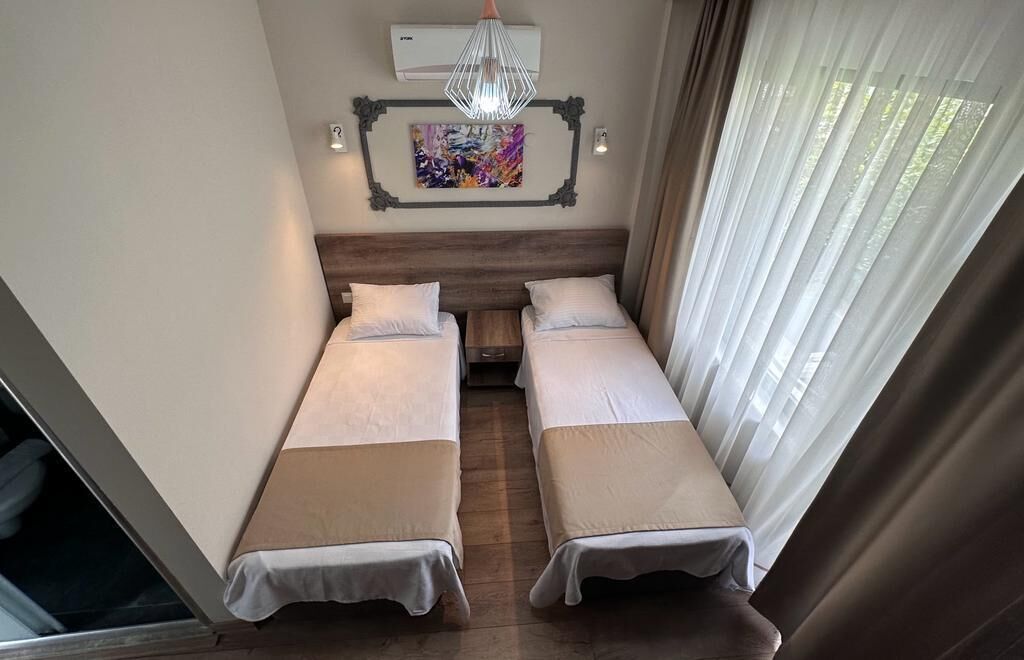 A room with two single beds, a wooden headboard, a ceiling fan, an air conditioning unit, a colorful abstract painting, and a window with sheer curtains.