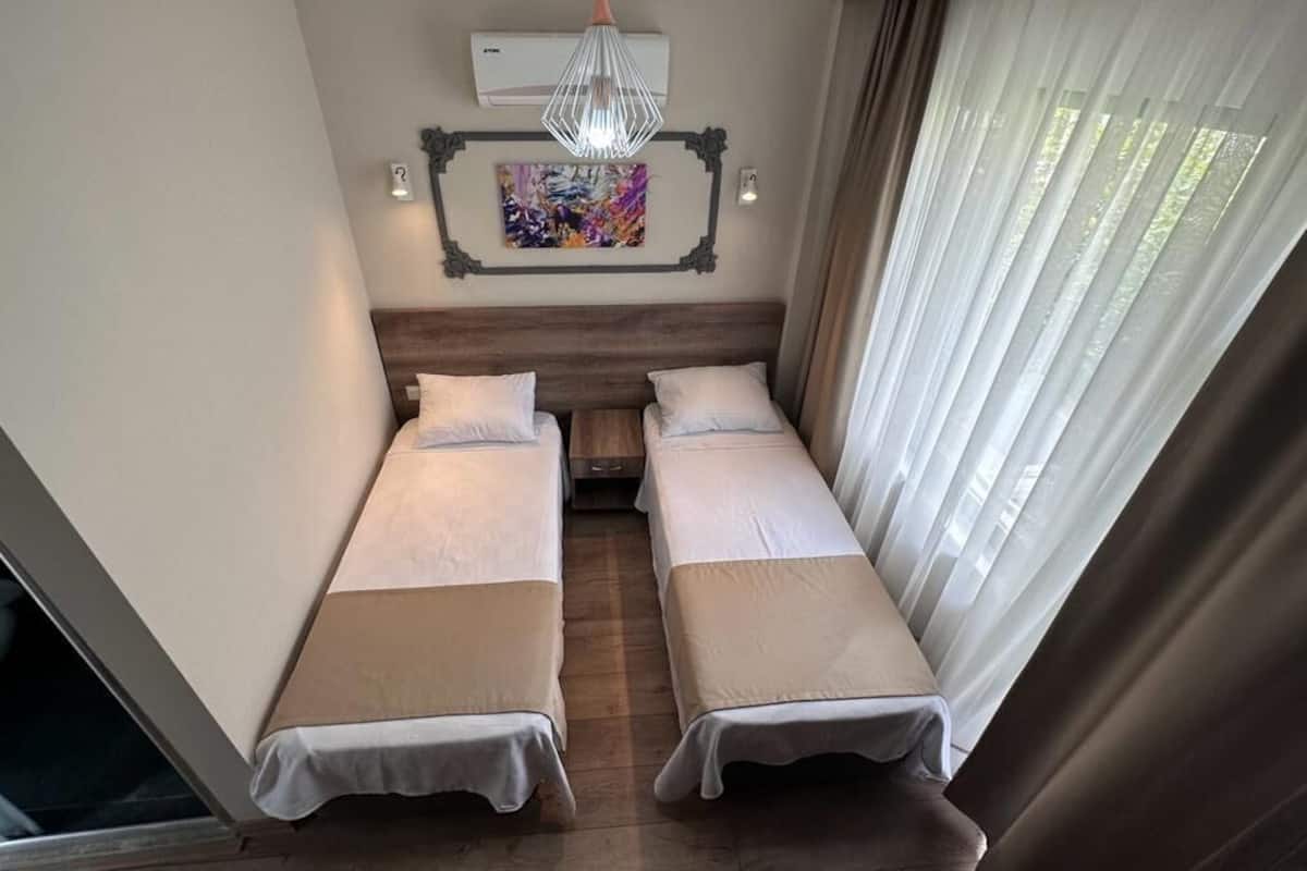 Une chambre avec deux lits simples, une tête de lit en bois, un ventilateur de plafond, un appareil de climatisation, un tableau abstrait aux couleurs vives et une fenêtre avec des rideaux légers.