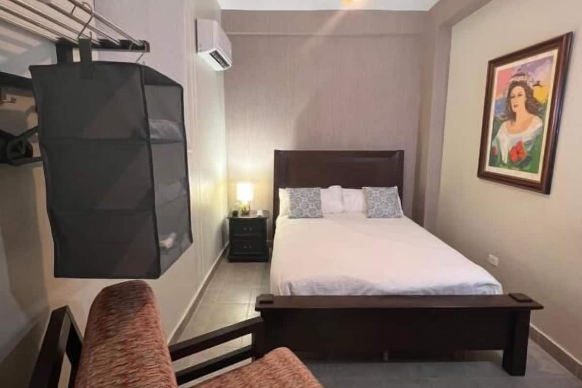 Een slaapkamer met een bed, een stoel, een nachtkastje, een lamp en een schilderij aan de muur.